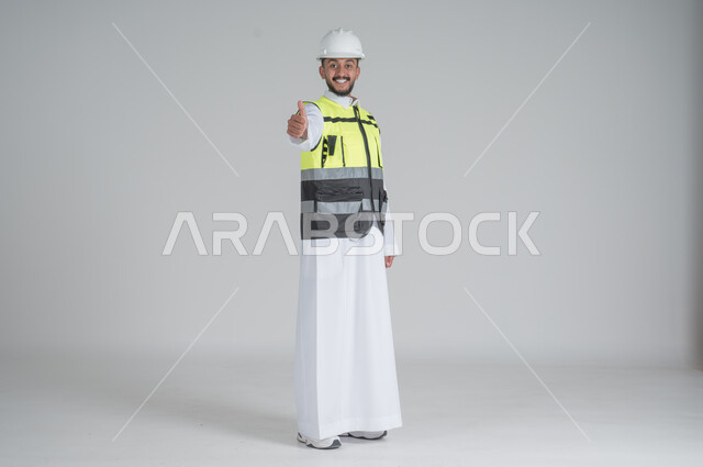 رفع اليد بايماءات الاعجاب والقبول, النظر الى الكاميرا بتعابير السعادة والسرور, مهن ووظائف سعودية, تنمية وتطوير المجال الهندسي, بورتريه لرجل عربي سعودي خليجي يرتدي الثوب والسترة ويضع خوذه الحماية, صورة الجسم بالطول الكامل, خلفية بيضاء