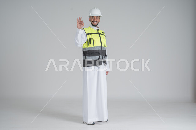 رفع اليد بايماءات الاعجاب والقبول, النظر الى الكاميرا بتعابير السعادة والسرور, مهن ووظائف سعودية, تنمية وتطوير المجال الهندسي, بورتريه لرجل عربي سعودي خليجي يرتدي الثوب والسترة ويضع خوذه الحماية, صورة الجسم بالطول الكامل, خلفية بيضاء