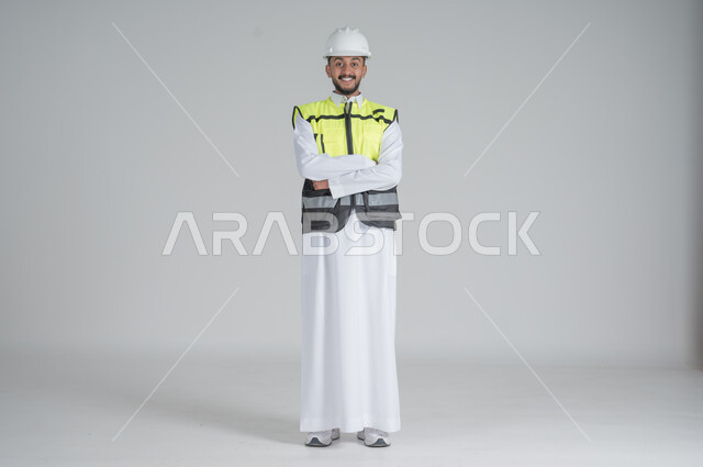 الوقوف بايدي متقاطعة بتعابير السعادة والسرور, مهن ووظائف سعودية, بورتريه لرجل عربي سعودي خليجي يرتدي الثوب والسترة ويضع خوذه الحماية, صورة الجسم بالطول الكامل, خلفية بيضاء