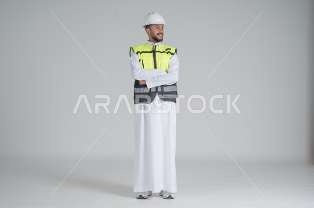 الوقوف بايدي متقاطعة بتعابير السعادة والسرور, مهن ووظائف سعودية, بورتريه لرجل عربي سعودي خليجي يرتدي الثوب والسترة ويضع خوذه الحماية, صورة الجسم بالطول الكامل, خلفية بيضاء