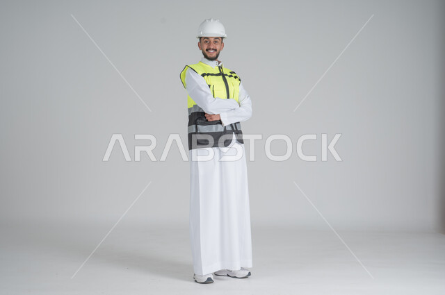 Standing with crossed hands with expressions of happiness and pleasure, Saudi professions and jobs, portrait of a Saudi Gulf Arab man wearing a thobe and jacket and wearing a protective helmet, full-length body portrait, white background