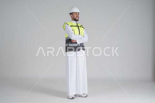 الوقوف بايدي متقاطعة بتعابير السعادة والسرور, مهن ووظائف سعودية, بورتريه لرجل عربي سعودي خليجي يرتدي الثوب والسترة ويضع خوذه الحماية, صورة الجسم بالطول الكامل, خلفية بيضاء