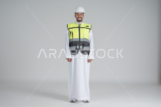 الوقوف باستقامة وثقة بالنفس, ايماءات السعادة والسرور, مهن ووظائف سعودية, بورتريه لرجل عربي سعودي خليجي يرتدي الثوب والسترة ويضع خوذه الحماية, صورة الجسم بالطول الكامل, خلفية بيضاء