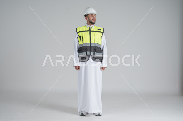 الوقوف باستقامة وثقة بالنفس, ايماءات السعادة والسرور, مهن ووظائف سعودية, بورتريه لرجل عربي سعودي خليجي يرتدي الثوب والسترة ويضع خوذه الحماية, صورة الجسم بالطول الكامل, خلفية بيضاء