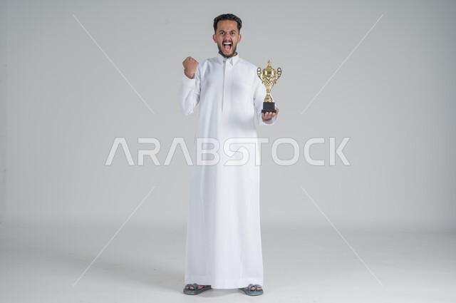 إيماءات وجه تدل على الشعور بالفخر والسعادة بالفوز، تشجيع الفرق والمنتخبات والاندية, بورتريه لشاب عربي خليجي سعودي يرتدي الثوب التقليدي ويحمل بيده كأس البطولة, صورة الجسم بالطول الكامل, خلفية بيضاء