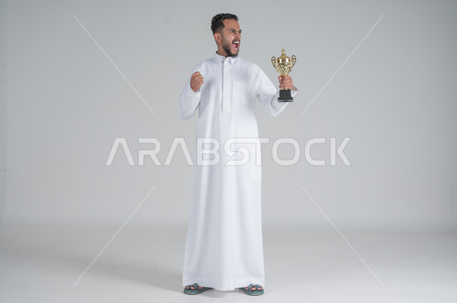إيماءات وجه تدل على الشعور بالفخر والسعادة بالفوز، تشجيع الفرق والمنتخبات والاندية, بورتريه لشاب عربي خليجي سعودي يرتدي الثوب التقليدي ويحمل بيده كأس البطولة, صورة الجسم بالطول الكامل, خلفية بيضاء