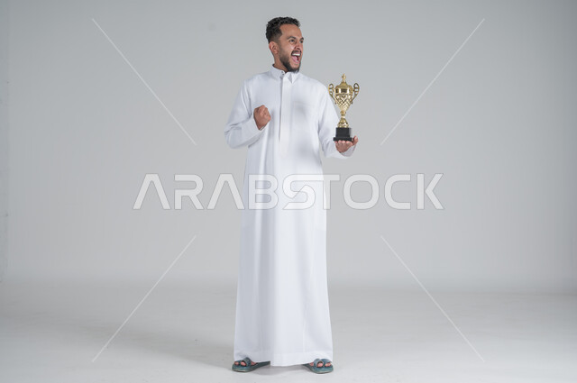 إيماءات وجه تدل على الشعور بالفخر والسعادة بالفوز، تشجيع الفرق والمنتخبات والاندية, بورتريه لشاب عربي خليجي سعودي يرتدي الثوب التقليدي ويحمل بيده كأس البطولة, صورة الجسم بالطول الكامل, خلفية بيضاء