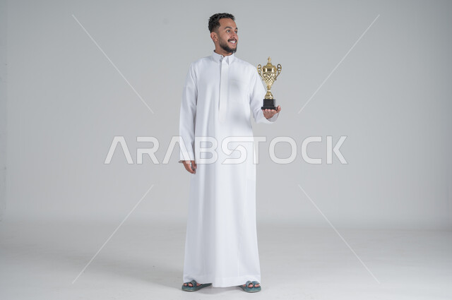 إيماءات وجه تدل على الشعور بالفخر والسعادة بالفوز، تشجيع الفرق والمنتخبات والاندية, بورتريه لشاب عربي خليجي سعودي يرتدي الثوب التقليدي ويحمل بيده كأس البطولة, صورة الجسم بالطول الكامل, خلفية بيضاء
