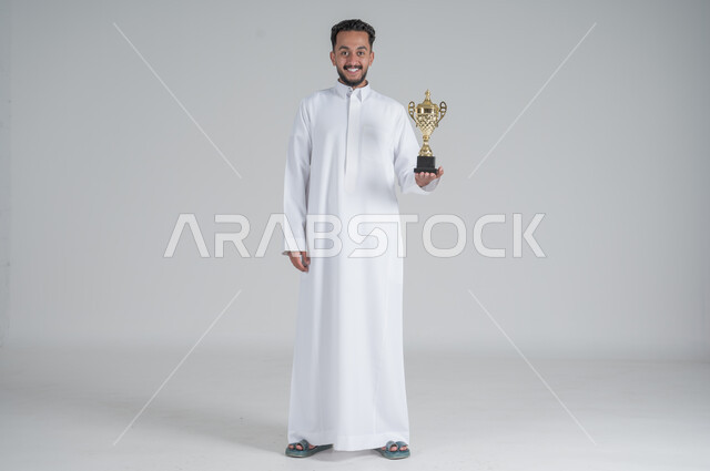 إيماءات وجه تدل على الشعور بالفخر والسعادة بالفوز، تشجيع الفرق والمنتخبات والاندية, بورتريه لشاب عربي خليجي سعودي يرتدي الثوب التقليدي ويحمل بيده كأس البطولة, صورة الجسم بالطول الكامل, خلفية بيضاء