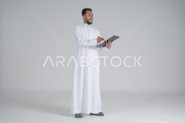 تعابير السعادة والسرور, تدوين و تسجيل الملاحظات, بورتريه لرجل عربي سعودي خليجي يرتدي الثوب التقليدي و يمسك بيديه مفكرة وقلم يقوم بكتابة المعلومات, صورة الجسم بالطول الكامل, خلفيه بيضاء