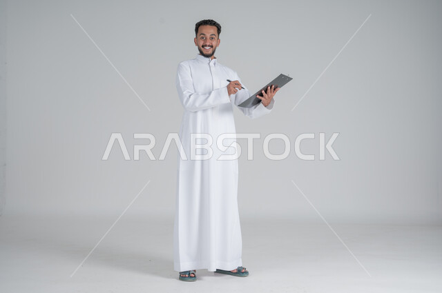 تعابير السعادة والسرور, تدوين و تسجيل الملاحظات, بورتريه لرجل عربي سعودي خليجي يرتدي الثوب التقليدي و يمسك بيديه مفكرة وقلم يقوم بكتابة المعلومات, صورة الجسم بالطول الكامل, خلفيه بيضاء