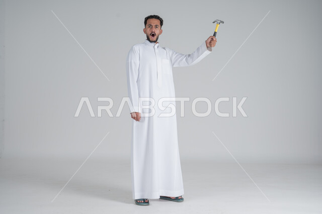 Gestures of shock and wonder, skill and mastery of work, using maintenance tools and equipment to carry out repairs, portrait of a Saudi Gulf Arab man wearing a traditional dress and holding a hammer in his hand, full-length body portrait, white background.