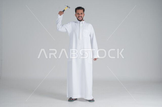 ايماءات القوة والجدية, المهارة و اتقان العمل، استخدام أدوات ومعدات الصيانة لاجراء التصليحات، بورتريه لرجل عربي سعودي خليجي يرتدي الثوب التقليدي ويرفع بيده مطرقة، صورة الجسم بالطول الكامل، خلفية بيضاء