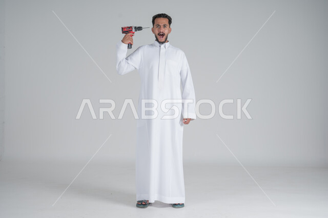 ايماءات الصدمة والترقب, المهارة و اتقان العمل، استخدام أدوات ومعدات الصيانة لاجراء التصليحات، بورتريه لرجل عربي سعودي خليجي يرتدي الثوب التقليدي ويحمل بيده فرد ثاقب يوجهه نحو رأسه، صورة الجسم بالطول الكامل، خلفية بيضاء