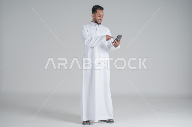 The concept of facilitating business and the ability to complete tasks, using modern technologies and advanced devices, portrait of a Saudi Gulf Arab young man wearing a traditional dress holding a calculator in his hand to perform calculations, full-length body, white background