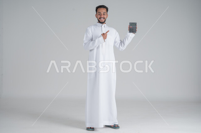 The concept of facilitating business and the ability to complete tasks, using modern technologies and advanced devices, portrait of a Saudi Gulf Arab young man wearing a traditional dress holding a calculator in his hand to perform calculations, full-length body, white background
