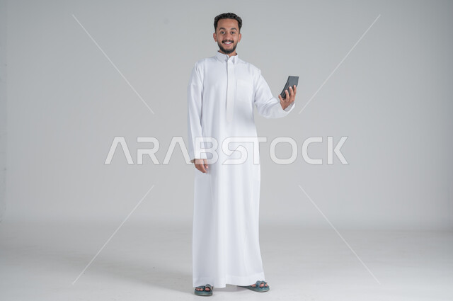 مفهوم تسهيل الاعمال والقدرة على إنجاز المهام، استخدام التقنيات الحديثة والأجهزة المتطورة ، بورتريه لشاب عربي خليجي سعودي يرتدي الثوب التقليدي يمسك بيده آلة حاسبة لإجراء عمليات الحساب، الجسم بالطول الكامل، خلفية بيضاء