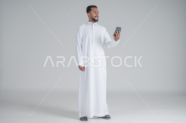 مفهوم تسهيل الاعمال والقدرة على إنجاز المهام، استخدام التقنيات الحديثة والأجهزة المتطورة ، بورتريه لشاب عربي خليجي سعودي يرتدي الثوب التقليدي يمسك بيده آلة حاسبة لإجراء عمليات الحساب، الجسم بالطول الكامل، خلفية بيضاء