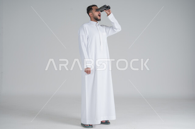 رؤية الاشياء البعيدة عبر المنظار، حب المغامرة والاستكشاف، بورتريه لشاب عربي خليجي سعودي يرتدي الثوب التقليدي وينظر من خلال المنظور بإيماءات وجه مختلفة, التخطيط المستقبلي للأهداف والطموحات، صورة الجسم بالطول الكامل، خلفية بيضاء