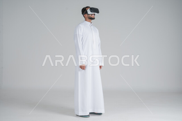 Technological and technical development, portrait of a Saudi Gulf Arab man wearing a traditional thobe and 3D virtual reality glasses, merging modern technologies with fun and entertainment, gestures of integration and concentration, full-length body image, white background