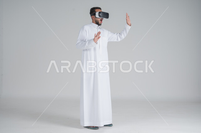 التطور التكنولوجي والتقني، بورتريه لرجل عربي خليجي سعودي يرتدي الثوب التقليدي ونظارات الواقع الافتراضي ثلاثية الأبعاد, دمج التقنيات الحديثة مع المتعة والتسلية، ايماءات الاندماج والتركيز، صورة الجسم بالطول الكامل، خلفية بيضاء
