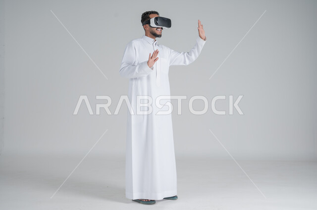 التطور التكنولوجي والتقني، بورتريه لرجل عربي خليجي سعودي يرتدي الثوب التقليدي ونظارات الواقع الافتراضي ثلاثية الأبعاد, دمج التقنيات الحديثة مع المتعة والتسلية، ايماءات الاندماج والتركيز، صورة الجسم بالطول الكامل، خلفية بيضاء