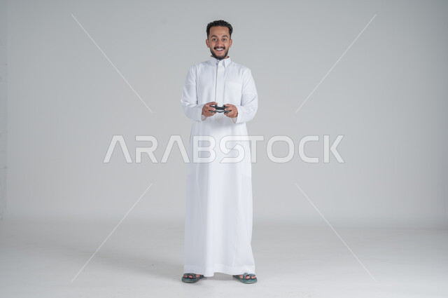 تقنيات الكترونية ترفيهيه, بورتريه لرجل عربي سعودي خليجي يرتدي الثوب التقليدي ويحمل بيده عصا التحكم، تجربة ممتعة ومثيرة في الأجهزة الحديثة، ايماءات السعادة والسرور، التفاعل والاندماج مع ألعاب البلايستيشن، صورة الجسم بالطول الكامل, خلفية بيضاء