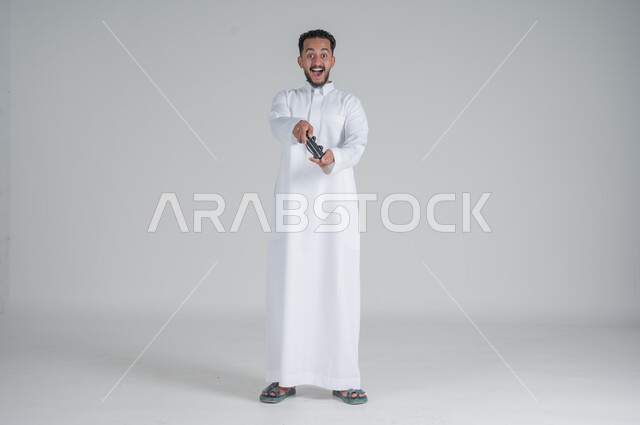 تقنيات الكترونية ترفيهيه, بورتريه لرجل عربي سعودي خليجي يرتدي الثوب التقليدي ويحمل بيده عصا التحكم، تجربة ممتعة ومثيرة في الأجهزة الحديثة، ايماءات السعادة والسرور، التفاعل والاندماج مع ألعاب البلايستيشن، صورة الجسم بالطول الكامل, خلفية بيضاء