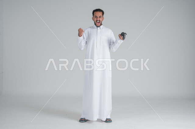 تقنيات الكترونية ترفيهيه, بورتريه لرجل عربي سعودي خليجي يرتدي الثوب التقليدي ويحمل بيده عصا التحكم، تجربة ممتعة ومثيرة في الأجهزة الحديثة، ايماءات السعادة والسرور، التفاعل والاندماج مع ألعاب البلايستيشن، صورة الجسم بالطول الكامل, خلفية بيضاء