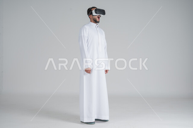 التطور التكنولوجي والتقني، بورتريه لرجل عربي خليجي سعودي يرتدي الثوب التقليدي ونظارات الواقع الافتراضي ثلاثية الأبعاد, دمج التقنيات الحديثة مع المتعة والتسلية، ايماءات الاندماج والصدمة، صورة الجسم بالطول الكامل، خلفية بيضاء