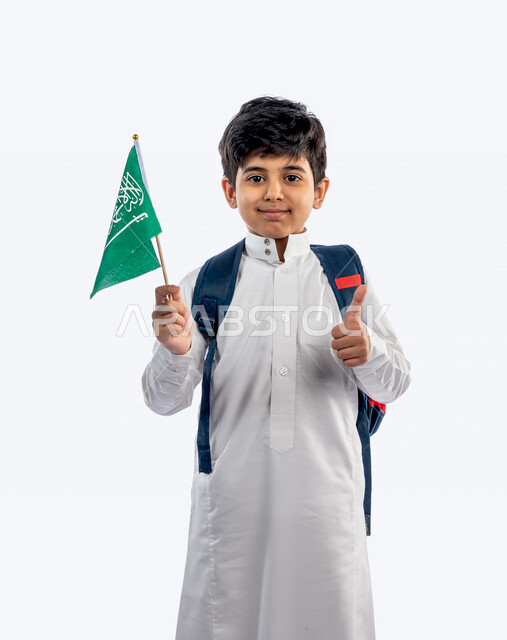 بورتريه لتلميذ مدرسي عربي خليجي سعودي ، يرتدي حقيبة ظهر مدرسية ، و يرتدي الثوب السعودي التقليدي ، يحمل بيده علم السعودية و اليد الأخرى اشارة اعجاب ، ايماءات و تعابير للوجه و اليدين تدل على السعادة ، عام دراسي جديد ، مدرسة سعودية