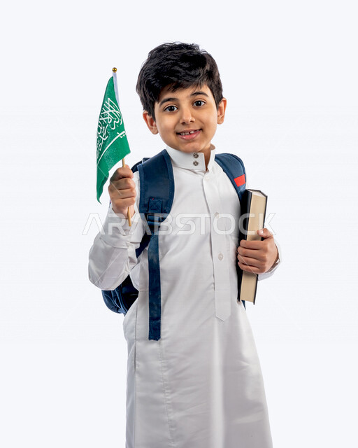 Portrait of a Saudi Arabian Gulf school student, wearing a school backpack, and wearing the traditional Saudi thobe, holding the book and the Saudi flag in his hand, gestures and facial expressions and hands indicating happiness, new school year, Saudi sc