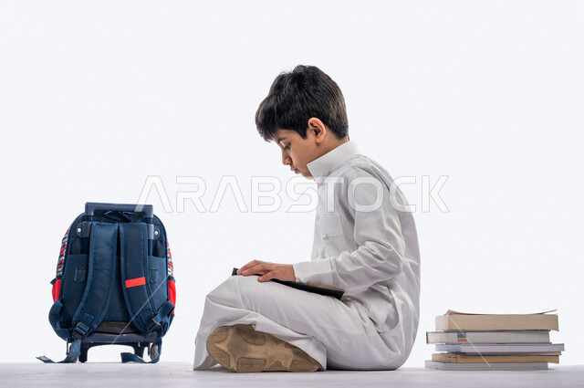بورتريه لتلميذ مدرسي عربي خليجي سعودي مبتسم ، يجلس بجانب حقيبة ظهر مدرسية و مجموعة من الكتب المدرسية ، يرتدي الثوب السعودي التقليدي ، بيده كتاب مفتوح يقرأ به ، ايماءات و تعابير وجه و يدين مختلفة ، عام دراسي جديد