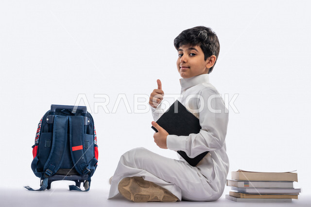 بورتريه لتلميذ مدرسي عربي خليجي سعودي مبتسم ، يجلس بجانب حقيبة ظهر مدرسية و مجموعة من الكتب ، يرتدي الثوب السعودي التقليدي ، يرفع أصبع الابهام لعمل اشارة اعجاب ، ايماءات و تعابير وجه و يدين مختلفة ، عام دراسي جديد ، الاستمتاع بقراءة الكتب المدرسية