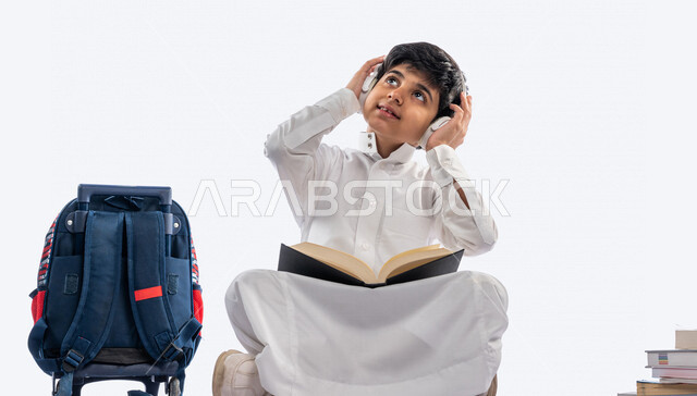 بورتريه لتلميذ مدرسي عربي خليجي سعودي مبتسم ، يجلس بجانب حقيبة ظهر مدرسية ، يرتدي الثوب السعودي التقليدي  و سماعة رأس ، ايماءات و تعابير وجه و يدين مختلفةتدل علة السعادة ، عام دراسي جديد ، الاستمتاع بقراءة الكتاب المدرسي