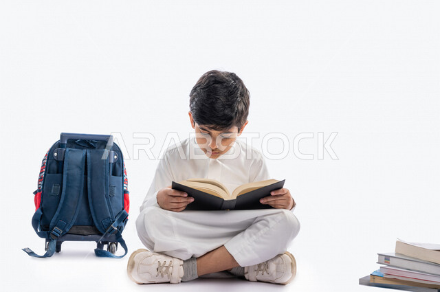 بورتريه لتلميذ مدرسي عربي خليجي سعودي مبتسم ، يجلس بجانب حقيبة ظهر مدرسية و مجموعة من الكتب المدرسية ، يرتدي الثوب السعودي التقليدي ، بيده كتاب مفتوح يقرأ به ، ايماءات و تعابير وجه و يدين مختلفة ، عام دراسي جديد ، الاستمتاع بقراءة الكتاب المدرسي