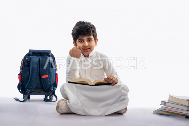 بورتريه لتلميذ مدرسي عربي خليجي سعودي مبتسم ، يجلس بجانب حقيبة ظهر مدرسية و مجموعة من الكتب المدرسية ، يرتدي الثوب السعودي التقليدي ، بيده كتاب مفتوح يقرأ به ، ايماءات و تعابير وجه و يدين مختلفة  تدل على الجد و الاجتهاد و القوة ، عام دراسي جديد