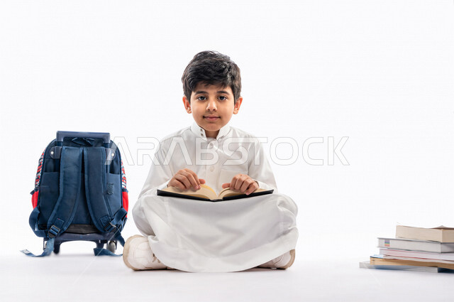 بورتريه لتلميذ مدرسي عربي خليجي سعودي مبتسم ، يجلس بجانب حقيبة ظهر مدرسية و مجموعة من الكتب المدرسية ، يرتدي الثوب السعودي التقليدي ، بيده كتاب مفتوح يقرأ به ، ايماءات و تعابير وجه و يدين مختلفة ، عام دراسي جديد ، الاستمتاع بقراءة الكتاب المدرسي ، مدرسة س