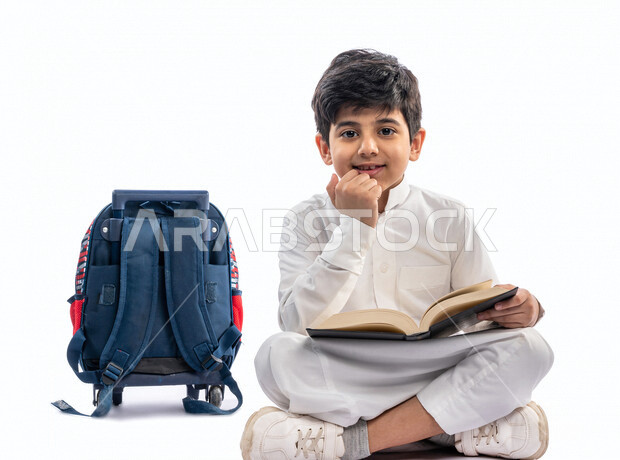 بورتريه لتلميذ مدرسي عربي خليجي سعودي مبتسم ، يجلس بجانب حقيبة ظهر مدرسية ، يرتدي الثوب السعودي التقليدي ، بيده كتاب مفتوح يقرأ به ، ايماءات و تعابير وجه و يدين مختلفة ، عام دراسي جديد ، الاستمتاع بقراءة الكتاب المدرسي ، مدرسة سعودية