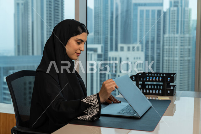 مفهوم إدارة المشاريع عن بعد، صورة مقربة لامرأة عربية خليجية اماراتية محجبة ترتدي عباءة سوداء تجلس في المكتب تقوم بانجاز مهام العمل عبر جهاز الحاسوب المحمول، شابة سعودية تقوم باستخدام الاجهزة التقنية الحديثة