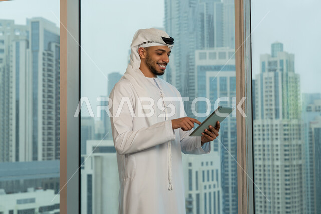 The concept of remote project management, an Arab Gulf Emirati man wearing a kandura and a ghutra stands in front of a glass facade overlooking the city, completing work tasks via a tablet, using modern technical devices, professionalism and dedication to achievement