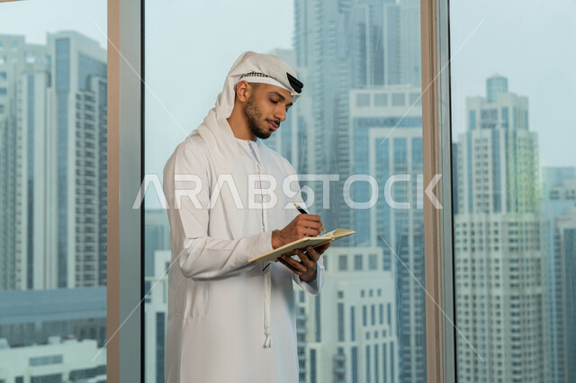 الاحترافية والتفاني في الإنجاز، ملفات ووثائق خاصة بالعمل، رجل عربي خليجي اماراتي يرتدي الكندورة والغترة يقف مقابل واجهة زجاجية مطلة على المدينة يقوم بتدوين وكتابة الملاحظات على الورق، دراسه وتتبع المشاريع، انجاز المهام وتحقيق الاهداف، مهن ووظائف مكتبيه