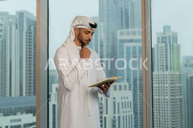 ملفات ووثائق خاصة بالعمل، رجل عربي خليجي اماراتي يرتدي الكندورة والغترة يقف مقابل واجهة زجاجية مطلة على المدينة يقوم بتدوين وكتابة الملاحظات على الورق، الاحترافية والتفاني في الإنجاز، دراسه وتتبع المشاريع، انجاز المهام وتحقيق الاهداف، مهن ووظائف مكتبيه