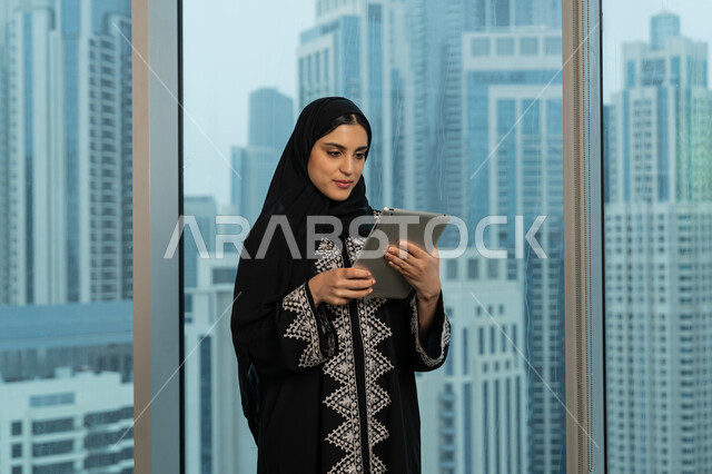 مفهوم إدارة المشاريع عن بعد، صورة مقربة لامرأة عربية خليجية اماراتية محجبة ترتدي عباءة سوداء تقف مقابل واجهة زجاجية مطلة على المدينة تقوم بانجاز مهام العمل عبر جهاز التابلت، شابة سعودية تقوم باستخدام الاجهزة التقنية الحديثة