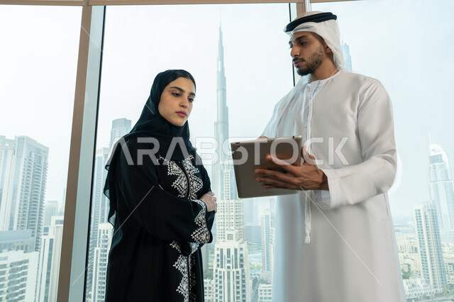 استخدام الجهاز اللوحي لتوضيح العمليات المتبعة، التفاهم والتعاون بين الزملاء لتحقيق أهداف الشركة، الاحترافية والتفاني في الإنجاز، رجل عربي خليجي اماراتي يرتدي الكندورة والغترة البيضاء يحمل بيده التابلت يقوم بشرح خطة العمل لزميلته، ضمان فهم واضح وشامل للاعمال