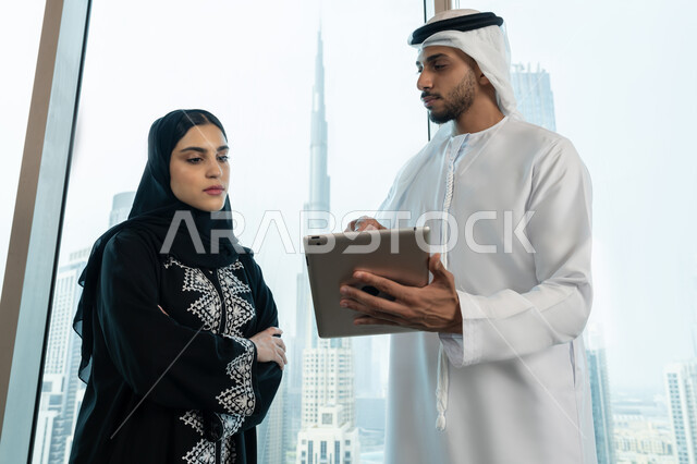 Professionalism and dedication to achievement, using the tablet to explain the processes followed, understanding and cooperation between colleagues to achieve the company's goals, an Arab Gulf Emirati man wearing a white kandura and ghutra holding a tablet in his hand explaining the work plan to his colleague, ensuring a clear and comprehensive understanding of the business