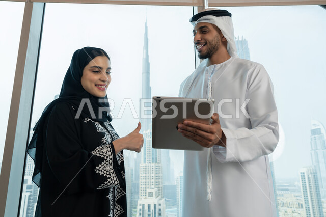 ايماءات الموافقة والقبول، ضمان فهم واضح وشامل للاعمال، الاحترافية والتفاني في الإنجاز، التفاهم والتعاون بين الزملاء لتحقيق أهداف الشركة، رجل عربي خليجي اماراتي يرتدي الكندورة والغترة البيضاء يحمل بيده التابلت يقوم بشرح خطة العمل لزميلته