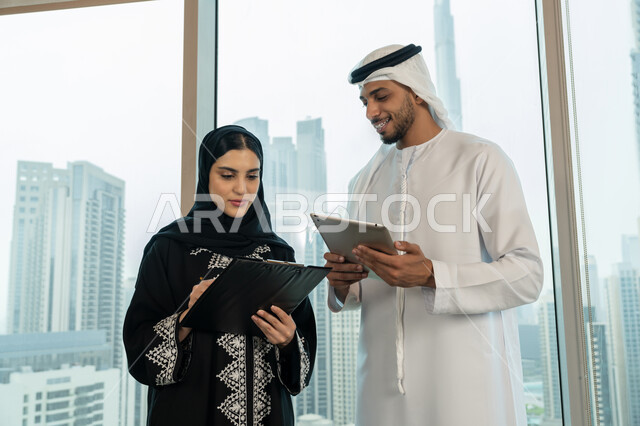 التحقق والتدقيق في العروض المالية، تطوير خطة وآلية العمل، كتابة وتسجيل البيانات والملاحظات، إنجاز الأعمال المكتبية، التفاهم والتعاون بين الزملاء لتحقيق أهداف الشركة، صورة مقربة لرجل عربي خليجي اماراتي يرتدي الكندورة والغترة يحمل التابلت يقوم بشرح المهام لزميلته