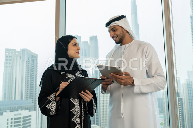 Developing a work plan and mechanism, verifying and auditing financial offers, writing and recording data and notes, completing office work, understanding and cooperation between colleagues to achieve the company's goals, a close-up image of an Emirati Gulf Arab man wearing a kandura and a ghutra holding a tablet explaining tasks to his colleague