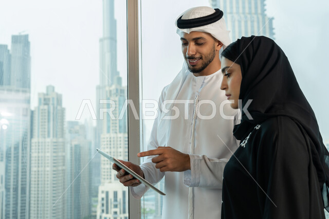 Using the tablet to explain the processes followed, understanding and cooperation between colleagues to achieve the company's goals, professionalism and dedication to achievement, an Arab Gulf Emirati man wearing a white kandura and ghutra holding a tablet in his hand explaining the work plan to his colleague, ensuring a clear and comprehensive understanding of the work